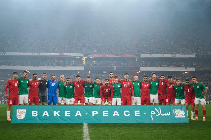 Group of male football players in green jerseys with numbers and names stand side by side with players in red jerseys on a green soccer field under stadium lights with a crowd blurred in the background. They hold a large horizontal banner across the front reading BAKE BAKEAEA PEACE in white letters on green background with Arabic text below. Players appear diverse in age and build wearing standard athletic uniforms and cleats.
