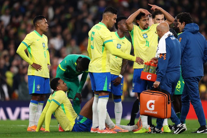 First image shows Brazilian national team players in yellow and green kits gathered on the soccer field during a match, with one player kneeling injured on the ground, medical staff in white and blue attire providing assistance, holding a Gatorade bottle and orange medical bag, stadium crowd in background. Second image depicts Gabriel Magalhaes in Brazilian kit standing near the goalpost, appearing distressed with hand on thigh, coach in blue jacket nearby, focus on apparent leg injury area.