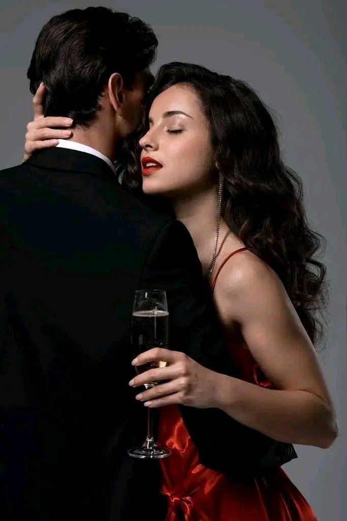 A man in a black suit embraces a woman from behind, his arms around her shoulders as he kisses her cheek, while she holds a champagne glass in her right hand and gazes slightly upward with red lips. The woman wears a red strapless dress with wavy dark hair cascading over her shoulders. The background is a neutral gray studio setting.