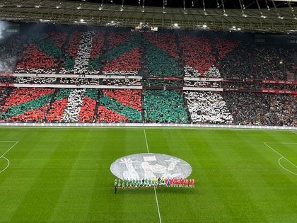 A large soccer stadium filled with a massive crowd holding up green white and red flags and banners forming a giant mosaic pattern across the stands behind the goal line with smoke or fog on the field and a central circular emblem on the green grass pitch players lined up in formation near the center circle under bright stadium lights