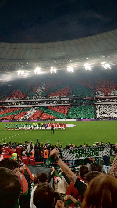 Stadium at night with bright lights illuminating the field and stands filled with spectators in green white and red colors forming a flag pattern across sections players in red and green uniforms standing in formation on the green grass a large banner held by fans reading Euskal Herria Askatu in the foreground crowds cheering with raised arms and flags