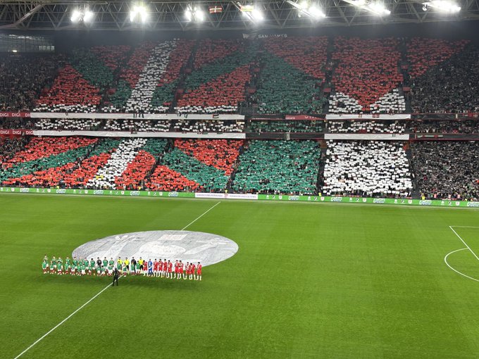 A large soccer stadium with stands packed with spectators holding and waving green white and red Basque flags mixed with black white and green Palestinian flags creating a colorful mosaic across multiple sections. The field below features a green pitch with a white center circle and players from two teams standing in formation one team in green jerseys and shorts the other in red with white accents all facing the crowd under illuminated floodlights. The atmosphere conveys unity and national pride through the flag displays.