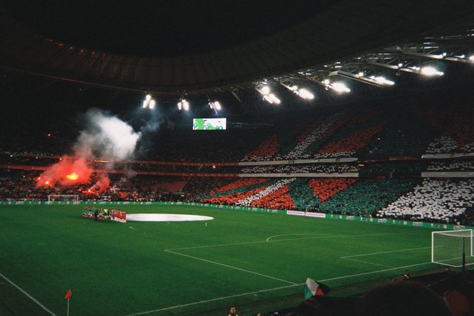 Nighttime view of a large soccer stadium packed with fans in the stands holding up flags and banners in red white and green stripes creating a colorful striped pattern across multiple sections with thick white smoke rising from flares and pyrotechnics on the field side illuminating the area in bright red and green glows the green grass field visible in the center with white goalposts and a white circle marking at one end overhead stadium lights shining down and a covered upper tier in the background