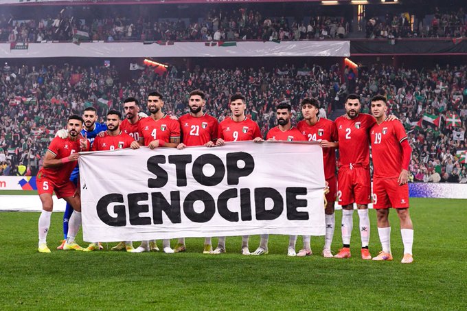 A group of male soccer players wearing red jerseys with numbers and white shorts stand in a line on a green grass field in a large stadium filled with spectators. They hold a large white banner displaying the words STOP GENOCIDE in bold black capital letters. A Palestinian flag is visible in the background among the crowd. The players appear to be from a professional team based on their uniforms.