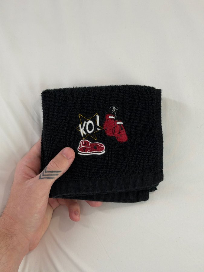 Black hand towel folded in thirds, held in a person’s left hand. The towel has embroidered red boxing gloves, a red boxing shoe, and a yellow starburst with the letters “KO!” on the front.