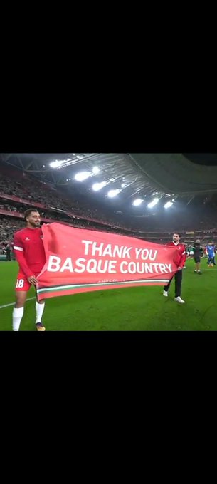 Two individuals dressed in red soccer uniforms with number 18 visible on one stand on a green grass field holding a large red banner that reads Thank you Basque Country in white letters. The background shows a large stadium interior with bright overhead lights illuminating the scene and a crowd of spectators seated in the stands. The setting appears to be during or after a professional soccer match.
