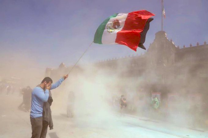 A man in a blue shirt and gray pants holds a pole with a large green white and red Mexican flag waving in the wind during a dusty outdoor event near ornate stone buildings with arches and other people standing around in casual clothing.