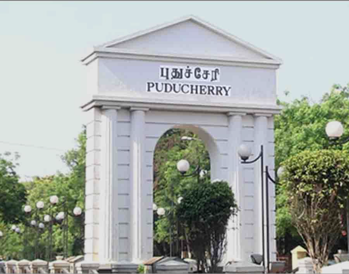 White neoclassical archway structure with Tamil and English text reading Puducherry flanked by green trees bushes and street lamps under clear sky