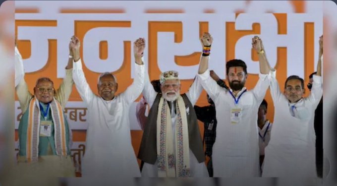Group of five men in traditional Indian attire including white kurtas shawls and turbans standing on stage raising hands in victory pose. Prominent central figure wears orange vest and tilak on forehead. Background features orange banner with Hindi text. Setting appears to be political rally with supporters.