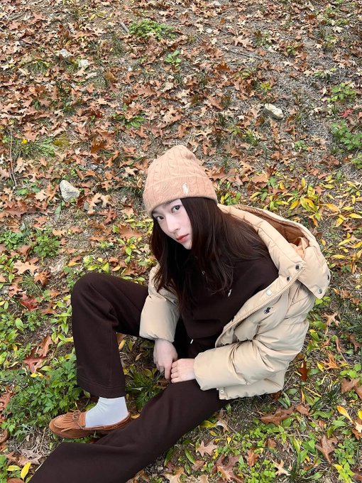 First image: A woman with long straight black hair and fair skin sits on grass covered in orange and yellow fallen leaves, wearing a light beige knit beanie, cream hooded puffer jacket with hood down, black pants, white crew socks, and brown loafers, smiling at camera with hands on knees. Background shows more scattered leaves and blurred trees. Second image: The same woman stands on a dirt path lined with tall trees bearing orange autumn foliage, wearing the same outfit, looking at camera with slight smile, hands in pockets. Third image: Close-up of the woman from low angle, hood up on cream puffer jacket, black shirt underneath, black pants, standing on path with blurred orange leaves and trees behind. Fourth image: The woman sits on grass with legs extended, wearing cream puffer jacket, black pants, white socks, brown loafers, long hair loose, gesturing with one hand, surrounded by orange fallen leaves and tree trunks in background.