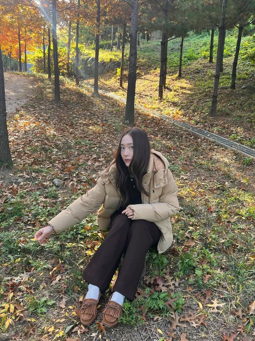 First image: A woman with long straight black hair and fair skin sits on grass covered in orange and yellow fallen leaves, wearing a light beige knit beanie, cream hooded puffer jacket with hood down, black pants, white crew socks, and brown loafers, smiling at camera with hands on knees. Background shows more scattered leaves and blurred trees. Second image: The same woman stands on a dirt path lined with tall trees bearing orange autumn foliage, wearing the same outfit, looking at camera with slight smile, hands in pockets. Third image: Close-up of the woman from low angle, hood up on cream puffer jacket, black shirt underneath, black pants, standing on path with blurred orange leaves and trees behind. Fourth image: The woman sits on grass with legs extended, wearing cream puffer jacket, black pants, white socks, brown loafers, long hair loose, gesturing with one hand, surrounded by orange fallen leaves and tree trunks in background.