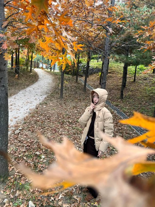 First image: A woman with long straight black hair and fair skin sits on grass covered in orange and yellow fallen leaves, wearing a light beige knit beanie, cream hooded puffer jacket with hood down, black pants, white crew socks, and brown loafers, smiling at camera with hands on knees. Background shows more scattered leaves and blurred trees. Second image: The same woman stands on a dirt path lined with tall trees bearing orange autumn foliage, wearing the same outfit, looking at camera with slight smile, hands in pockets. Third image: Close-up of the woman from low angle, hood up on cream puffer jacket, black shirt underneath, black pants, standing on path with blurred orange leaves and trees behind. Fourth image: The woman sits on grass with legs extended, wearing cream puffer jacket, black pants, white socks, brown loafers, long hair loose, gesturing with one hand, surrounded by orange fallen leaves and tree trunks in background.