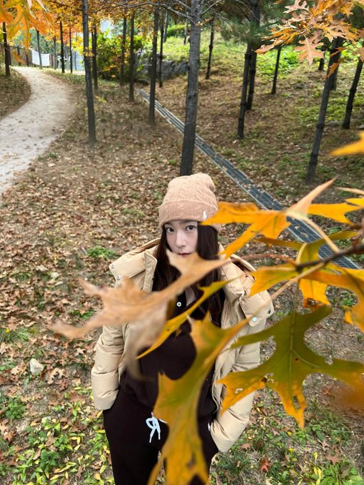First image: A woman with long straight black hair and fair skin sits on grass covered in orange and yellow fallen leaves, wearing a light beige knit beanie, cream hooded puffer jacket with hood down, black pants, white crew socks, and brown loafers, smiling at camera with hands on knees. Background shows more scattered leaves and blurred trees. Second image: The same woman stands on a dirt path lined with tall trees bearing orange autumn foliage, wearing the same outfit, looking at camera with slight smile, hands in pockets. Third image: Close-up of the woman from low angle, hood up on cream puffer jacket, black shirt underneath, black pants, standing on path with blurred orange leaves and trees behind. Fourth image: The woman sits on grass with legs extended, wearing cream puffer jacket, black pants, white socks, brown loafers, long hair loose, gesturing with one hand, surrounded by orange fallen leaves and tree trunks in background.