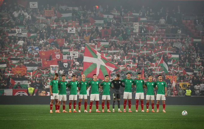 First image shows eleven male soccer players in green jerseys and white shorts standing in a line on green grass field with a soccer ball nearby goalkeeper in green kit others with red socks some with armbands stadium background filled with crowd holding green and white flags Danish flags and banners under overcast sky. Second image depicts soccer goalpost on green field with advertising boards for Kutxabank and BBVA stadium stands packed with enthusiastic fans waving red and white flags lighting red flares producing smoke crowd in various attire including green scarves.