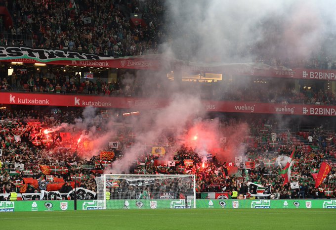 First image shows eleven male soccer players in green jerseys and white shorts standing in a line on green grass field with a soccer ball nearby goalkeeper in green kit others with red socks some with armbands stadium background filled with crowd holding green and white flags Danish flags and banners under overcast sky. Second image depicts soccer goalpost on green field with advertising boards for Kutxabank and BBVA stadium stands packed with enthusiastic fans waving red and white flags lighting red flares producing smoke crowd in various attire including green scarves.