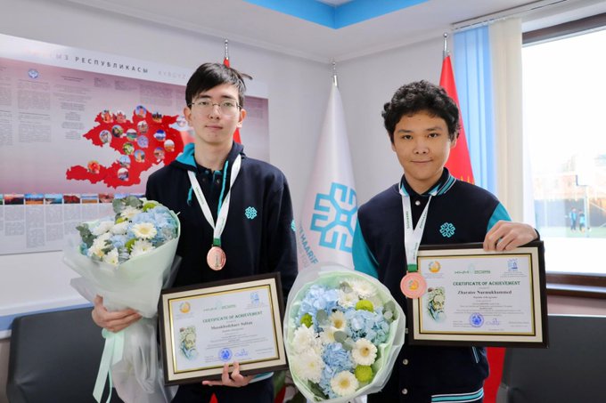 Two young male students in dark blue uniforms with school logos stand side by side in an indoor room with flags and educational posters on the walls. The student on the left, wearing glasses, holds a bouquet of white flowers and two framed certificates. The student on the right holds a bouquet of blue flowers, a gold-framed certificate, and a bronze medal on a ribbon around his neck. Both students smile and display their awards proudly.