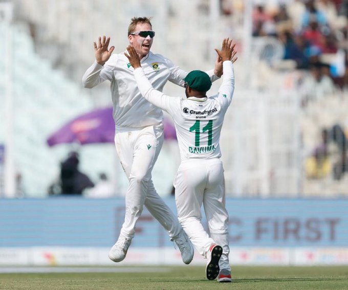 Two male cricketers in white uniforms celebrate a wicket on a green field at a stadium with purple umbrella and Grandstand sponsor visible one with number 11 on green cap and the other in sunglasses raising hands joyfully