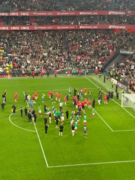 First image shows a large soccer stadium interior with a green field and circular center mark, surrounded by packed stands displaying a mosaic of green white red and orange flags forming patterns like crosses and stripes. Second image displays a stadium scoreboard reading 51 396 under Basque text for attendance, with illuminated sections and sponsor logos visible in the background. Third image captures the soccer field with players in red and green uniforms gathered near the center circle, referees in black, and stadium seating with green and red elements in the stands. Fourth image depicts a crowded urban street at dusk with historic buildings, a large group of people holding Palestinian flags, some wearing green hats, and others in casual attire waving banners.
