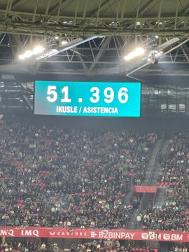 First image shows a large soccer stadium interior with a green field and circular center mark, surrounded by packed stands displaying a mosaic of green white red and orange flags forming patterns like crosses and stripes. Second image displays a stadium scoreboard reading 51 396 under Basque text for attendance, with illuminated sections and sponsor logos visible in the background. Third image captures the soccer field with players in red and green uniforms gathered near the center circle, referees in black, and stadium seating with green and red elements in the stands. Fourth image depicts a crowded urban street at dusk with historic buildings, a large group of people holding Palestinian flags, some wearing green hats, and others in casual attire waving banners.
