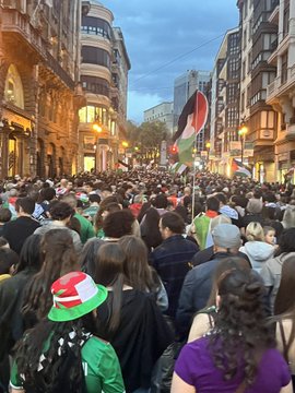 First image shows a large soccer stadium interior with a green field and circular center mark, surrounded by packed stands displaying a mosaic of green white red and orange flags forming patterns like crosses and stripes. Second image displays a stadium scoreboard reading 51 396 under Basque text for attendance, with illuminated sections and sponsor logos visible in the background. Third image captures the soccer field with players in red and green uniforms gathered near the center circle, referees in black, and stadium seating with green and red elements in the stands. Fourth image depicts a crowded urban street at dusk with historic buildings, a large group of people holding Palestinian flags, some wearing green hats, and others in casual attire waving banners.