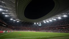 Night view of San Mames stadium exterior illuminated in red and white stripes with digital screen displaying match information and crowds outside. Lineup of players from both teams in red green and white jerseys standing on green field with Athletic Club banner behind and a red flag in air amid stadium seating. Player in red Palestine jersey number 4 raising arms in victory pose with coach nearby on field surrounded by other players in various jerseys and green turf. Wide night view of packed San Mames stadium interior with green field players and red seating filled with spectators under bright lights.