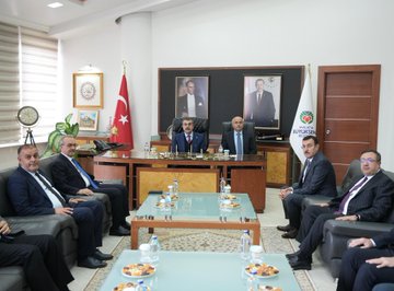 First image shows a young girl in a pink puffer vest and gray pants extending her hand while holding a large bouquet of white and red flowers wrapped in paper, facing a smiling middle-aged man in a dark blue suit, red tie, and white shirt who holds the bouquet and extends his hand, with other people in suits and a car visible in the background on a cloudy day. Second image depicts two men in dark suits and ties standing behind a white podium with gold accents and a logo, flanked by Turkish and white flags on stands, a screen showing a mans face, and lights on a plain wall. Third image portrays several men in dark suits seated around a glass-topped wooden table with water glasses and tea cups in a formal room with Turkish and green flags on walls, portraits, a clock, and plants.