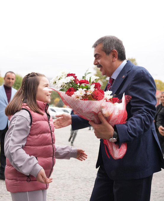 First image shows a young girl in a pink puffer vest and gray pants extending her hand while holding a large bouquet of white and red flowers wrapped in paper, facing a smiling middle-aged man in a dark blue suit, red tie, and white shirt who holds the bouquet and extends his hand, with other people in suits and a car visible in the background on a cloudy day. Second image depicts two men in dark suits and ties standing behind a white podium with gold accents and a logo, flanked by Turkish and white flags on stands, a screen showing a mans face, and lights on a plain wall. Third image portrays several men in dark suits seated around a glass-topped wooden table with water glasses and tea cups in a formal room with Turkish and green flags on walls, portraits, a clock, and plants.