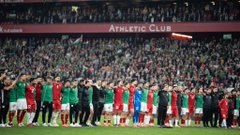 First image shows San Mames stadium at night illuminated in red and white stripes with a large screen displaying text, surrounded by crowds and lights. Second image depicts two teams of soccer players in green and red jerseys lined up on the field before a match, with a flag in the air and packed stands behind. Third image captures a player in a red Palestine jersey number 4 raising both arms in victory pose on the green field, surrounded by teammates and officials in various uniforms. Fourth image presents a wide night view of a full circular stadium packed with spectators, bright lights illuminating the green soccer field and surrounding areas.