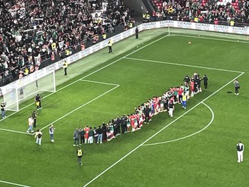 First image displays a full stadium view with the green pitch below and tiered stands filled with fans in sections of red green white and black colors forming patterns teams lined up in red and green jerseys around a large circular emblem on the field surrounded by sponsor banners. Second image captures the center circle on the lush green field with two teams of players in red and green uniforms standing in formation referees in black nearby and stadium seating packed with spectators under match organization banners. Third image shows a broad stadium interior with bright lights illuminating the green field where players in red jerseys gather near the goal line crowds filling stands in various colors including red and white. Fourth image focuses on the goal area with the net in the background green field extending out players from teams in red and green kits standing together near the corner flag referees and officials present with advertising boards along the sidelines and sparse crowd visible in the stands.