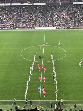 First image displays a full stadium view with the green pitch below and tiered stands filled with fans in sections of red green white and black colors forming patterns teams lined up in red and green jerseys around a large circular emblem on the field surrounded by sponsor banners. Second image captures the center circle on the lush green field with two teams of players in red and green uniforms standing in formation referees in black nearby and stadium seating packed with spectators under match organization banners. Third image shows a broad stadium interior with bright lights illuminating the green field where players in red jerseys gather near the goal line crowds filling stands in various colors including red and white. Fourth image focuses on the goal area with the net in the background green field extending out players from teams in red and green kits standing together near the corner flag referees and officials present with advertising boards along the sidelines and sparse crowd visible in the stands.