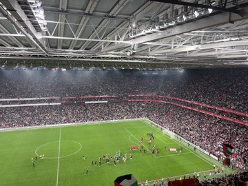 First image displays a full stadium view with the green pitch below and tiered stands filled with fans in sections of red green white and black colors forming patterns teams lined up in red and green jerseys around a large circular emblem on the field surrounded by sponsor banners. Second image captures the center circle on the lush green field with two teams of players in red and green uniforms standing in formation referees in black nearby and stadium seating packed with spectators under match organization banners. Third image shows a broad stadium interior with bright lights illuminating the green field where players in red jerseys gather near the goal line crowds filling stands in various colors including red and white. Fourth image focuses on the goal area with the net in the background green field extending out players from teams in red and green kits standing together near the corner flag referees and officials present with advertising boards along the sidelines and sparse crowd visible in the stands.