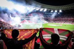 First image shows a large soccer stadium filled with spectators holding green, red, and white Basque flags creating a mosaic pattern across the stands, with smoke from red flares rising near the field and the green grass pitch visible below. Second image depicts two fans from behind, one holding a red and yellow flag and the other a green, red, and white Basque flag, overlooking a stadium field with smoke and bright lights in the background. Third image features the Osasuna soccer team in green jerseys and white shorts posing together on the field in front of a banner reading OFIZIALA TASUNA NA, with green and red scarves around their necks and the stadium stands behind. Fourth image shows the Iranian national soccer team in red jerseys holding a white banner that reads STOP GENOCIDE across it, standing on a green field with a blurred stadium crowd in the background.