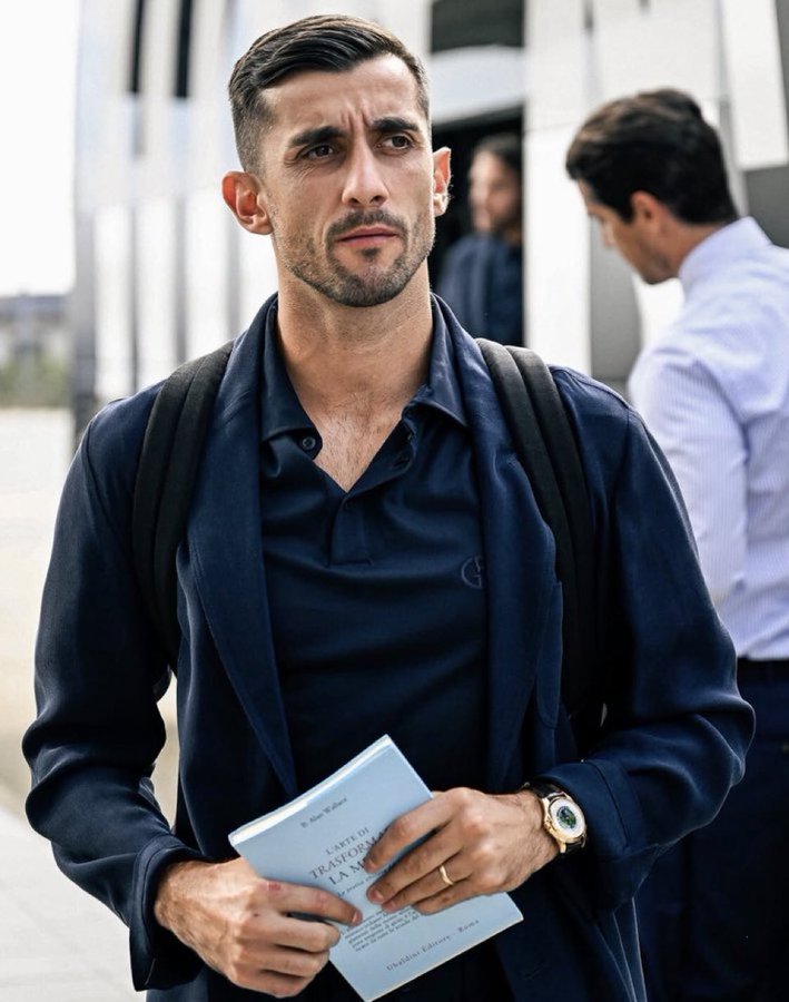 A man with short dark hair and a beard stands outdoors near a white building entrance wearing a dark blue polo shirt under a matching jacket and carrying a black backpack. He holds a white book titled with partial text visible in his right hand while wearing a gold wristwatch on his left wrist. Another man in a white shirt is visible in the background behind him and to the side.
