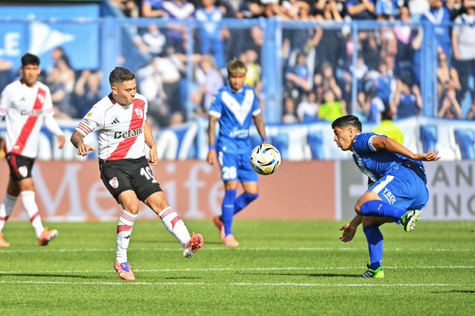 Two soccer players in red and white River Plate uniforms and blue Vélez Sarsfield uniforms compete for a ball mid-air on a green field with a crowd in the background behind a fence.