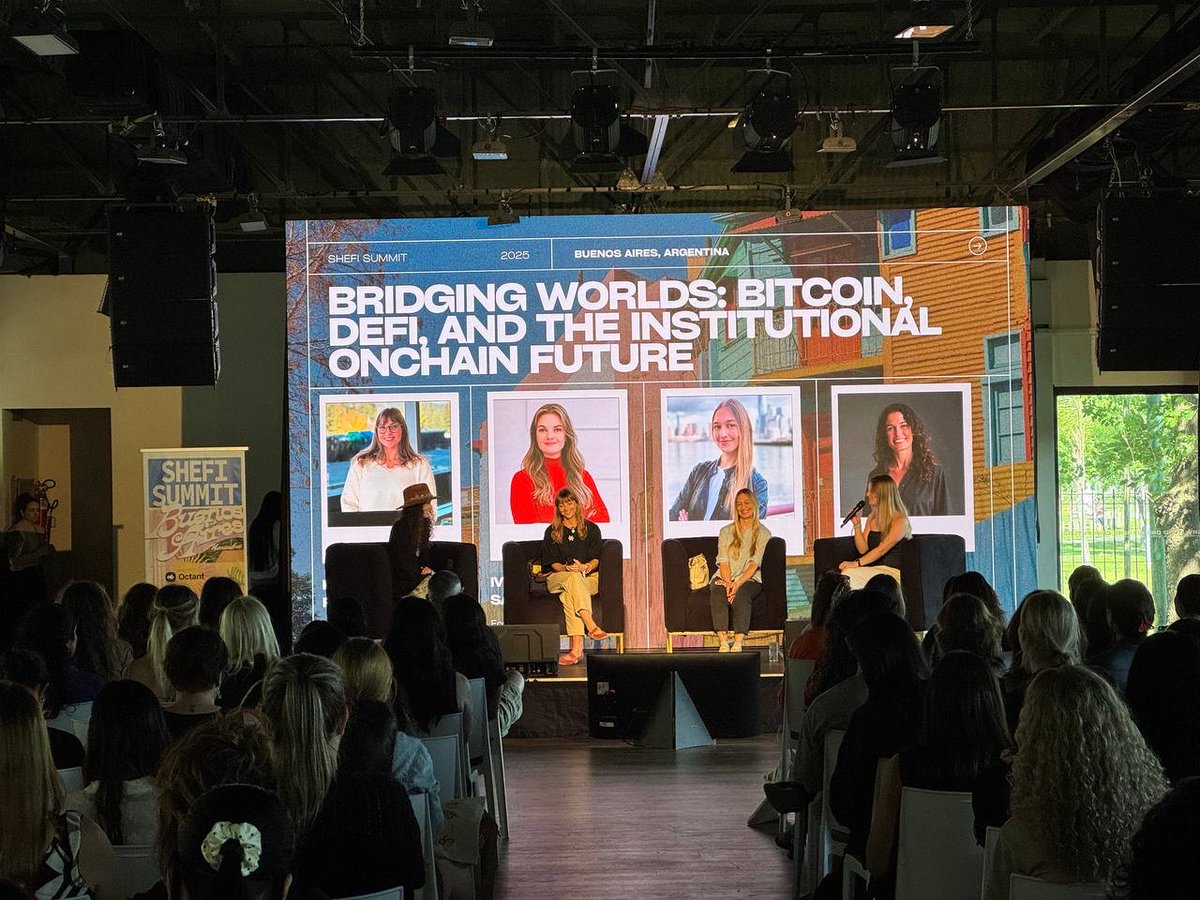 Panel of women speakers on stage at a SheFi summit discussing Bitcoin and DeFi, audience seated watching a large presentation screen.