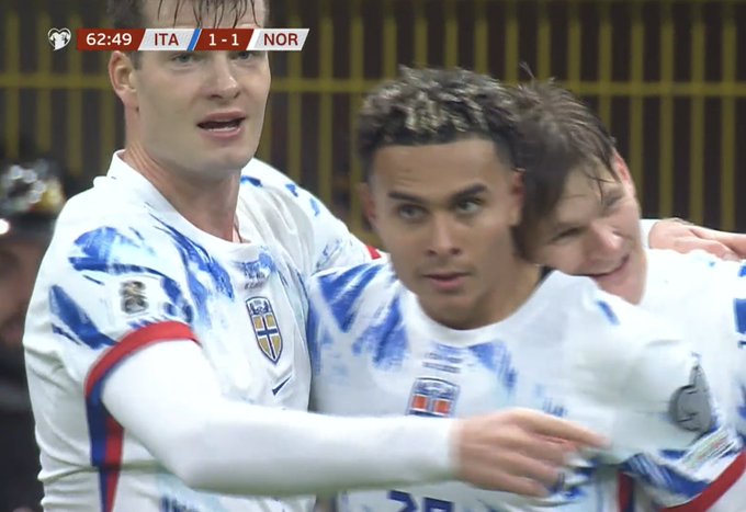 Three male football players in blue and white Norway national team uniforms with red accents stand close together on a green field, arms around each others shoulders, faces showing surprise and joy, one with wet hair, scoreboard overlay displays 62:49 time, ITA 1-1 NOR score, and match clock.