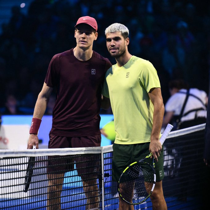 Two male tennis players stand side by side near a net on an indoor court with a crowd in the background. The player on the left wears a red cap, maroon shirt and shorts, wristband, and holds a racket in his right hand. The player on the right has platinum-dyed hair, wears a light green shirt and dark shorts, and holds a racket in his left hand. Both appear to be Jannik Sinner and Carlos Alcaraz based on the match context.