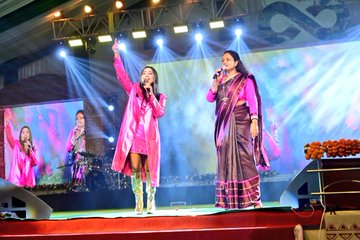 First image shows two women performing on a brightly lit stage: one in a pink shiny coat dress and gold boots holding a microphone, the other in a maroon saree with border also holding a microphone, with background musicians, drums, and stage lights. Second image depicts the two women embracing on stage under a banner with Bengali text and infinity symbol, surrounded by marigold flowers and stage setup. Third image features a woman in pink shiny coat dress and gold boots raising both arms on a red platform with blue backdrop and marigold decorations. Fourth image captures the two women holding hands on stage with sparklers bursting, one in pink outfit and the other in maroon saree, with a screen showing SH and stage elements.