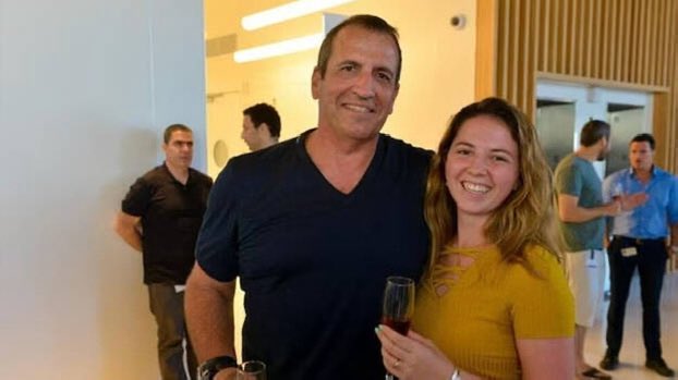 Two adults, a man in dark shirt holding a wine glass and a woman in yellow top smiling, stand closely in a modern indoor setting with beige walls, fluorescent lights, and blurred background of other men in casual attire at an event.