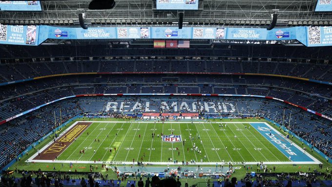 Aerial view of a large football stadium with green field marked for NFL play, surrounded by blue seats, banners displaying Real Madrid logo and NFL branding, empty stands, and overhead screens.