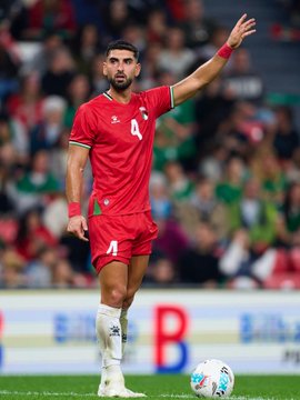 First image shows a male athlete in red soccer jersey number 4 standing on green field at packed stadium with blurred crowd, wearing white socks and cleats, arms at sides. Second image depicts the same athlete in red jersey and shorts number 4 raising right arm toward crowd, standing on field with soccer ball nearby and stadium seating in background. Third image captures two athletes mid-air competing for high ball, one in red uniform reaching up, the other in green uniform, surrounded by field and advertising banners. Fourth image features the red-uniformed athlete clapping hands near goalpost with net visible, another player in green uniform clapping in distance, stadium crowd and field in view.