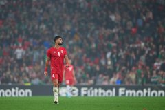 First image shows a male athlete in red soccer jersey number 4 standing on green field at packed stadium with blurred crowd, wearing white socks and cleats, arms at sides. Second image depicts the same athlete in red jersey and shorts number 4 raising right arm toward crowd, standing on field with soccer ball nearby and stadium seating in background. Third image captures two athletes mid-air competing for high ball, one in red uniform reaching up, the other in green uniform, surrounded by field and advertising banners. Fourth image features the red-uniformed athlete clapping hands near goalpost with net visible, another player in green uniform clapping in distance, stadium crowd and field in view.