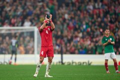First image shows a male athlete in red soccer jersey number 4 standing on green field at packed stadium with blurred crowd, wearing white socks and cleats, arms at sides. Second image depicts the same athlete in red jersey and shorts number 4 raising right arm toward crowd, standing on field with soccer ball nearby and stadium seating in background. Third image captures two athletes mid-air competing for high ball, one in red uniform reaching up, the other in green uniform, surrounded by field and advertising banners. Fourth image features the red-uniformed athlete clapping hands near goalpost with net visible, another player in green uniform clapping in distance, stadium crowd and field in view.