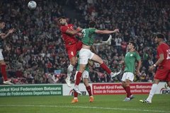 First image shows a male athlete in red soccer jersey number 4 standing on green field at packed stadium with blurred crowd, wearing white socks and cleats, arms at sides. Second image depicts the same athlete in red jersey and shorts number 4 raising right arm toward crowd, standing on field with soccer ball nearby and stadium seating in background. Third image captures two athletes mid-air competing for high ball, one in red uniform reaching up, the other in green uniform, surrounded by field and advertising banners. Fourth image features the red-uniformed athlete clapping hands near goalpost with net visible, another player in green uniform clapping in distance, stadium crowd and field in view.
