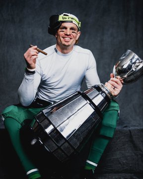 A man in a white long-sleeve shirt green football pants white socks black shoes and green headband with black eye blacking holds a lit cigar in one hand and a large silver trophy in the other smiling broadly while sitting on a dark surface with green confetti scattered around in a dimly lit studio setting. Similar pose in second image with trophy held closer and more confetti visible.