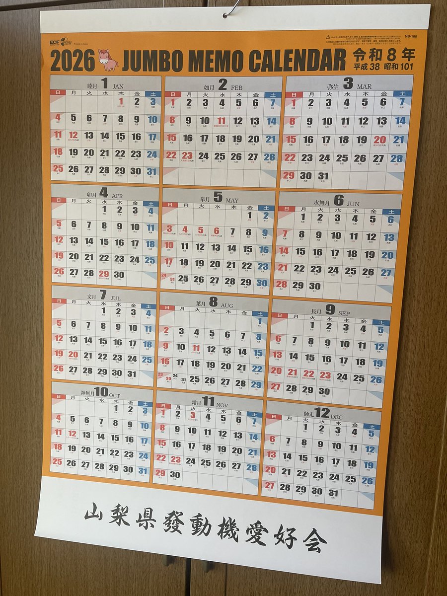 Large wall calendar for 2026 labeled Jumbo Memo Calendar in Japanese, with grid layouts for each month showing dates in black numbers on white and orange backgrounds, divided into sections for January to December, hung on a light wooden door or panel, and a white label at the bottom reading Yamanashi Hatsudōki Aikkai in Japanese characters.