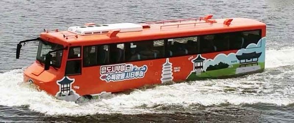 Orange amphibious bus with white roof and Korean-style pavilion decorations on sides travels on water surface, featuring text in Korean script indicating Baekma River tour service, surrounded by river waves and distant green landscape.