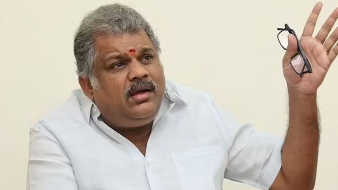Middle-aged Indian man with gray hair, mustache, wearing glasses in right hand, white short-sleeve shirt, gesturing with raised right hand, neutral background.