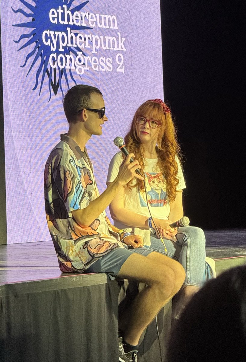 Vitalik Buterin sitting on stage at an Ethereum event wearing Moodeng shirt