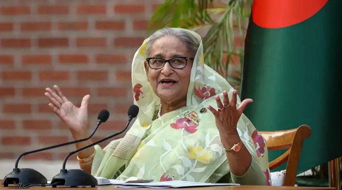 An elderly woman with glasses wearing a light green floral-patterned sari and matching headscarf stands at a wooden podium with two microphones. She gestures with both hands raised while speaking. A green Bangladesh flag is positioned to her right against a red brick wall with green plants. Papers and a watch are visible on the podium.